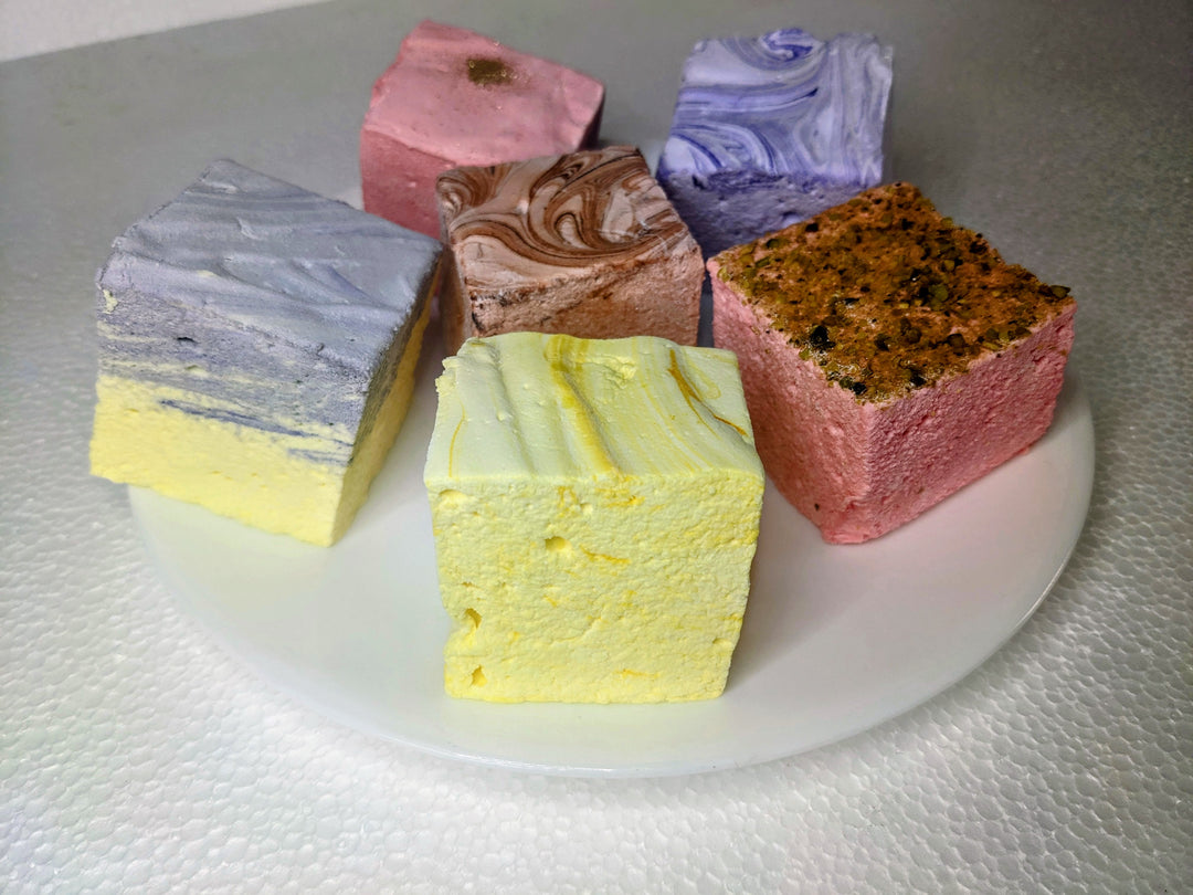 An elegant white plate displaying six varieties of gourmet marshmallows: lavender & honey, rose pistachio, creamy taro, champagne & sugar, lemon pound cake, and dulce de leche. Each marshmallow is handcrafted and generously sized