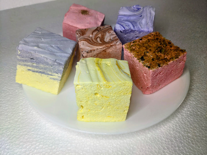 An elegant white plate displaying six varieties of gourmet marshmallows: lavender & honey, rose pistachio, creamy taro, champagne & sugar, lemon pound cake, and dulce de leche. Each marshmallow is handcrafted and generously sized