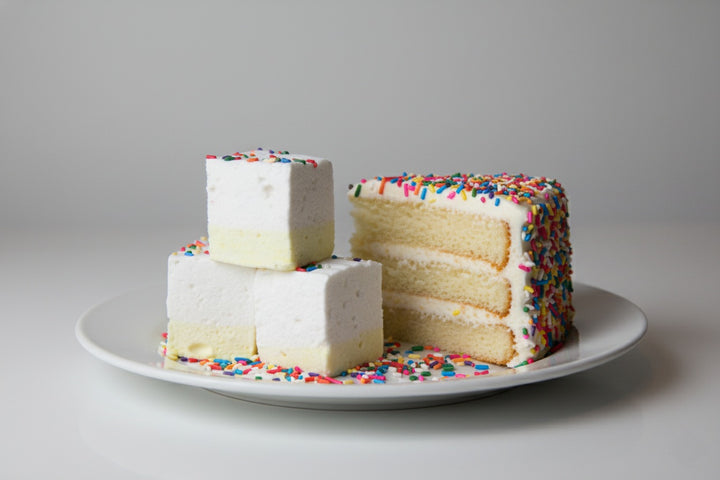 Three handcrafted birthday cake gourmet marshmallows made with beef gelatin and organic marshmallow root, featuring a white top, light yellow base, and rainbow sprinkles. Displayed on a white plate with a slice of sprinkle-topped birthday cake in the background. Clean-label, corn syrup-free marshmallows perfect for upscale gifting and summer-safe shipping.