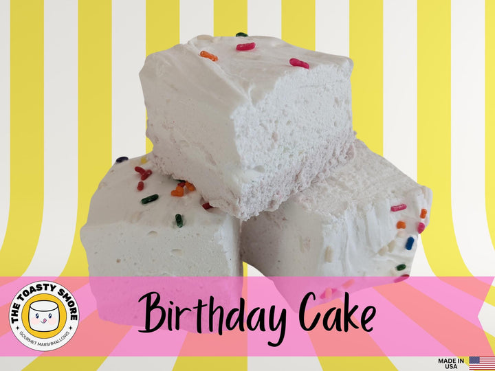 Birthday cake flavored marshmallows with a yellow bottom layer and white top layer, adorned with just a few colorful sprinkles. The marshmallows are displayed on a yellow and white striped background, featuring The Toasty Smore logo and a "Made in USA" flag.