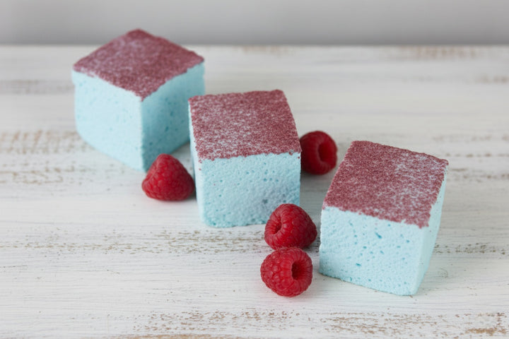Three blue raspberry gourmet marshmallows made with beef gelatin and organic marshmallow root, arranged in an angled line. Each marshmallow features a vibrant blue base with red sugar topping, styled alongside fresh red raspberries. Clean-label, corn syrup-free marshmallows designed for bold flavor and eye-catching presentation