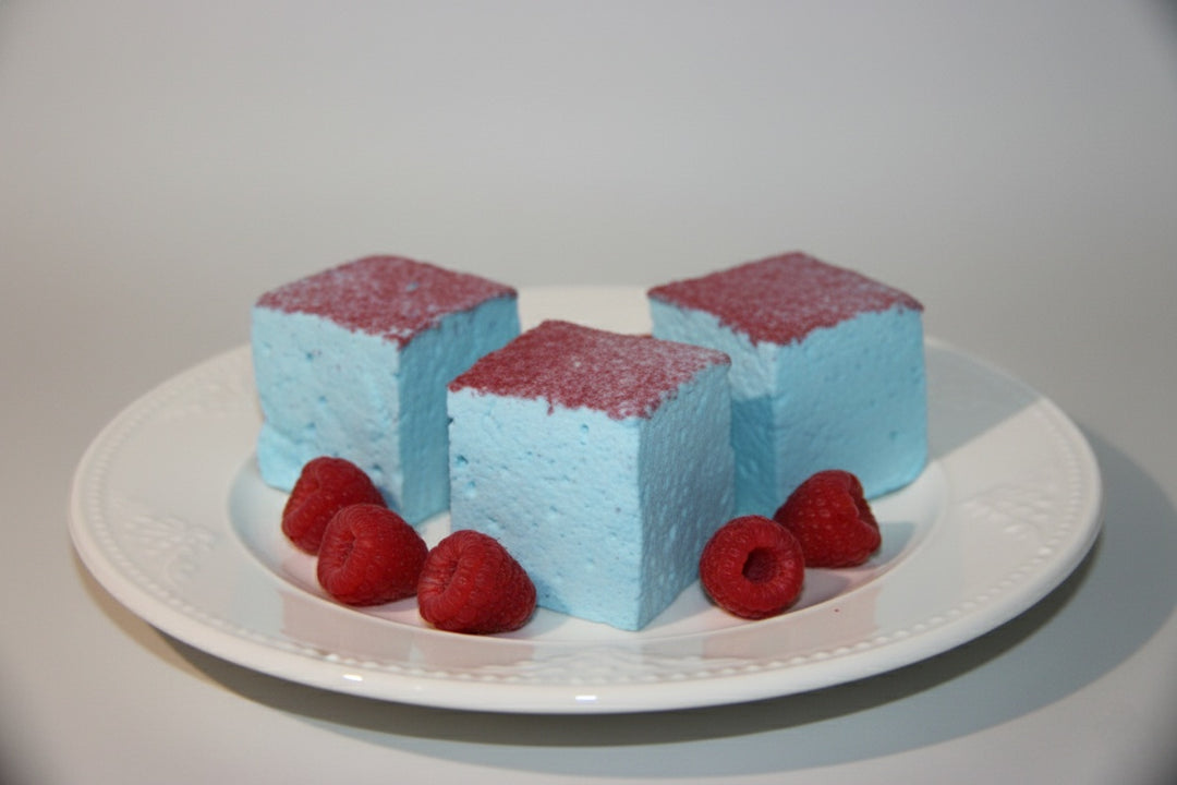 Three blue raspberry gourmet marshmallows made with beef gelatin and organic marshmallow root, topped with vibrant red sugar crystals. Arranged in a trio with fresh red raspberries in the foreground. Clean-label, corn syrup-free marshmallows crafted for bold flavor and eye-catching presentation.