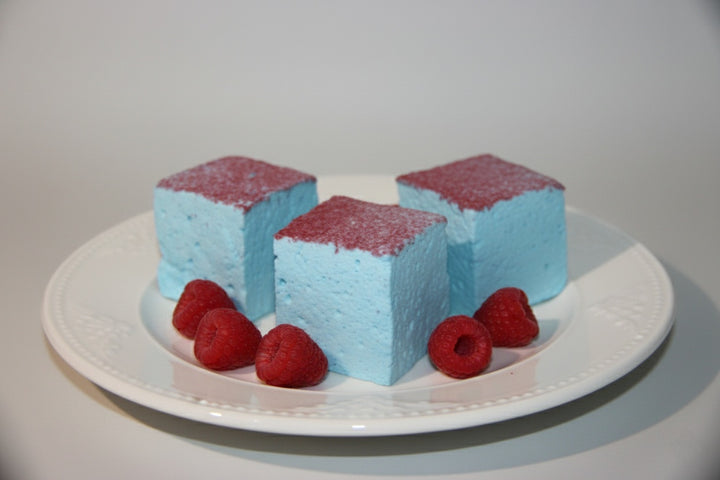 Three blue raspberry gourmet marshmallows made with beef gelatin and organic marshmallow root, topped with vibrant red sugar crystals. Arranged in a trio with fresh red raspberries in the foreground. Clean-label, corn syrup-free marshmallows crafted for bold flavor and eye-catching presentation.