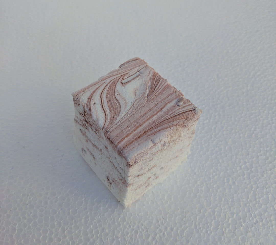 A close-up view of a single coffee-flavored gourmet marshmallow, handcrafted and made in the USA by The Toasty Smore, resting on a white plate. The marshmallow is white with rich brown swirls, creating a marbled effect that highlights the bold coffee infusion. Its soft, fluffy texture and intricate swirls emphasize its artisanal quality. Perfect for coffee lovers, this best gourmet marshmallow offers a delicious balance of sweetness and deep coffee flavor, ideal for pairing with hot drinks or enjoying as a 