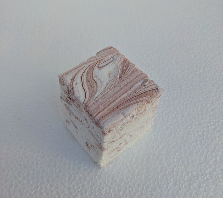 A close-up view of a single coffee-flavored gourmet marshmallow, handcrafted and made in the USA by The Toasty Smore, resting on a white plate. The marshmallow is white with rich brown swirls, creating a marbled effect that highlights the bold coffee infusion. Its soft, fluffy texture and intricate swirls emphasize its artisanal quality. Perfect for coffee lovers, this best gourmet marshmallow offers a delicious balance of sweetness and deep coffee flavor, ideal for pairing with hot drinks or enjoying as a 