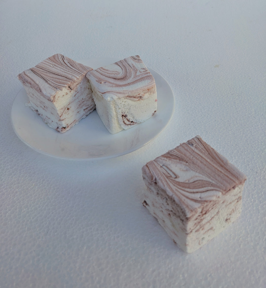 A close-up view of the best gourmet marshmallows, handcrafted and made in the USA by The Toasty Smore, arranged on a white plate with one marshmallow in front and two in the back. Each marshmallow is white with rich brown swirls, creating a marbled effect that highlights the bold coffee infusion. Their soft, fluffy texture and intricate swirls emphasize their artisanal quality. Perfect for coffee lovers, these marshmallows offer a delicious balance of sweetness and deep coffee flavor, ideal for pairing with