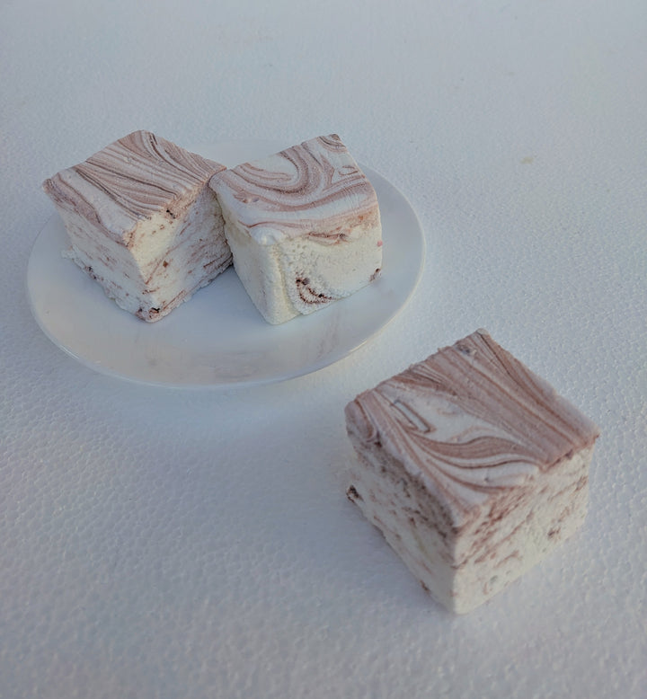 A close-up view of the best gourmet marshmallows, handcrafted and made in the USA by The Toasty Smore, arranged on a white plate with one marshmallow in front and two in the back. Each marshmallow is white with rich brown swirls, creating a marbled effect that highlights the bold coffee infusion. Their soft, fluffy texture and intricate swirls emphasize their artisanal quality. Perfect for coffee lovers, these marshmallows offer a delicious balance of sweetness and deep coffee flavor, ideal for pairing with