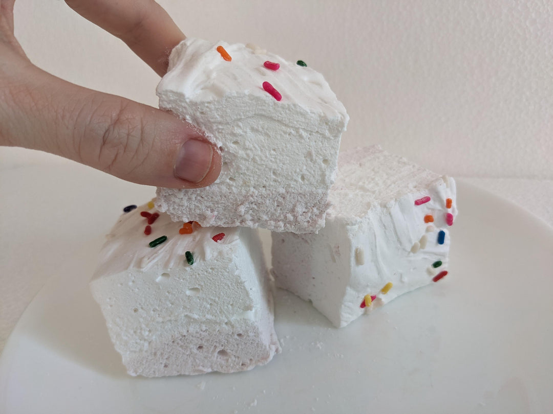 A hand picking up one birthday cake flavored marshmallow with a yellow bottom layer and white top layer, adorned with colorful sprinkles. The marshmallow is displayed on a clean, white background, featuring The Toasty Smore logo and a "Made in USA" flag.