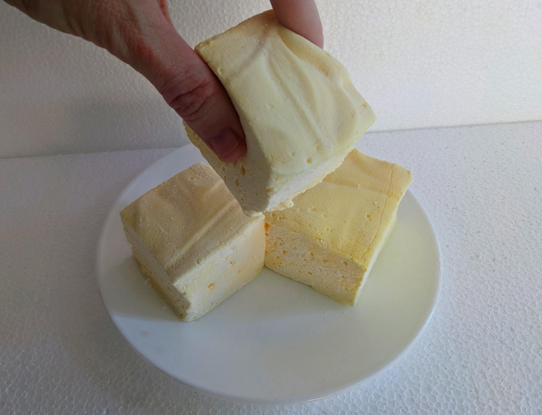 Three extra-large Butter Vanilla gourmet marshmallows made with organic marshmallow root and premium beef gelatin, displayed on a white plate against a plain white background. A hand is picking up one marshmallow.