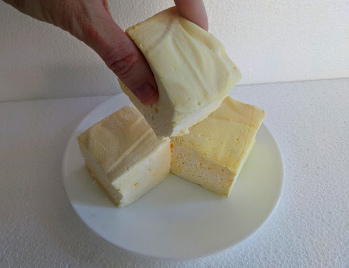 Three extra-large Butter Vanilla gourmet marshmallows made with organic marshmallow root and premium beef gelatin, displayed on a white plate against a plain white background. A hand is picking up one marshmallow.
