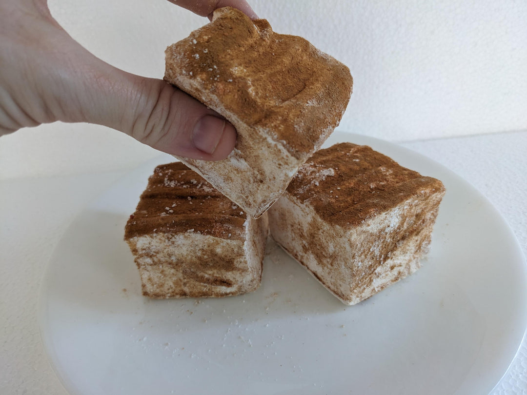 Three caramel churro marshmallows on a white plate, displayed on a plain white background. A hand is seen picking up one of the marshmallows.
