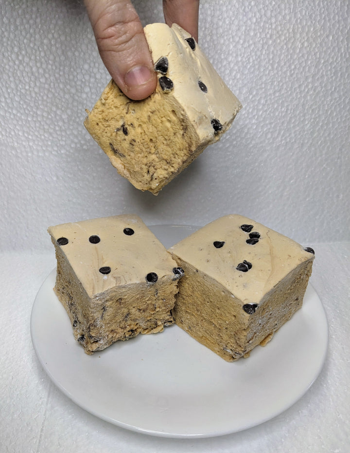 Three cookie dough marshmallows on a white plate, displayed on a clean, white background. A hand is seen picking up one of the marshmallows.