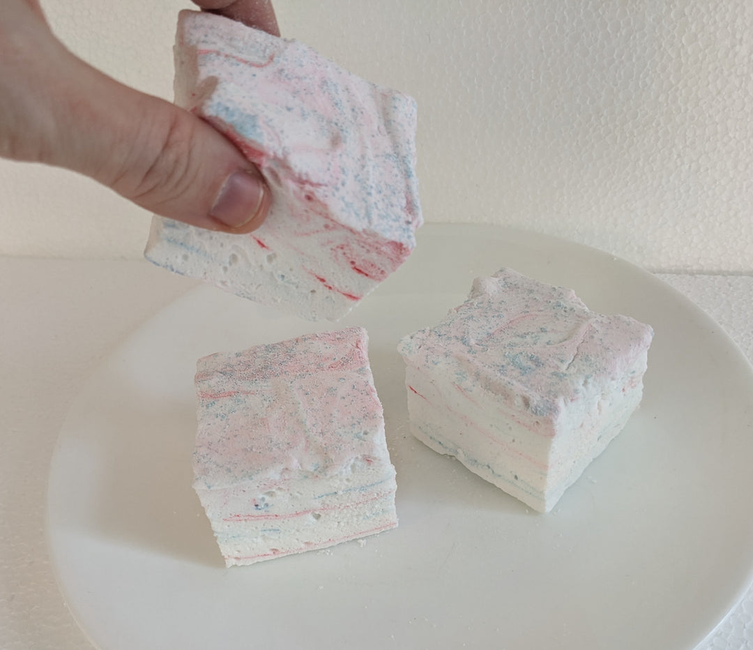 Three cotton candy marshmallows on a clean, white background. A hand is seen picking up one of the marshmallows.