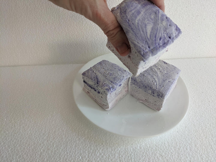 Three creamy purple taro marshmallows on a white plate, displayed on a plain white background. A hand is seen picking up one of the marshmallows.