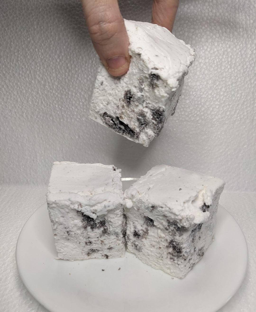 Three handcrafted gourmet cookies & cream marshmallows on a white plate, displayed on a plain white background. A hand is seen picking up one of the marshmallows. The marshmallows are approximately 2" x 2" x 2" each and made from premium gelatin.