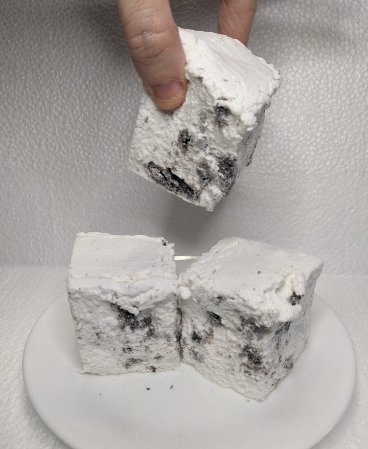 Three handcrafted gourmet cookies & cream marshmallows on a white plate, displayed on a plain white background. A hand is seen picking up one of the marshmallows. The marshmallows are approximately 2" x 2" x 2" each and made from premium gelatin.