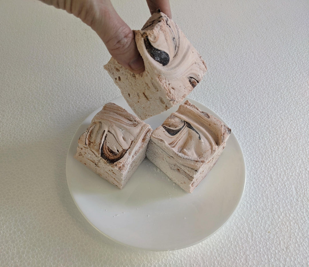 Three handcrafted gourmet Dulce De Leche marshmallows, neatly placed on a white plate and displayed against a plain white background. A hand is shown picking up one of the marshmallows.