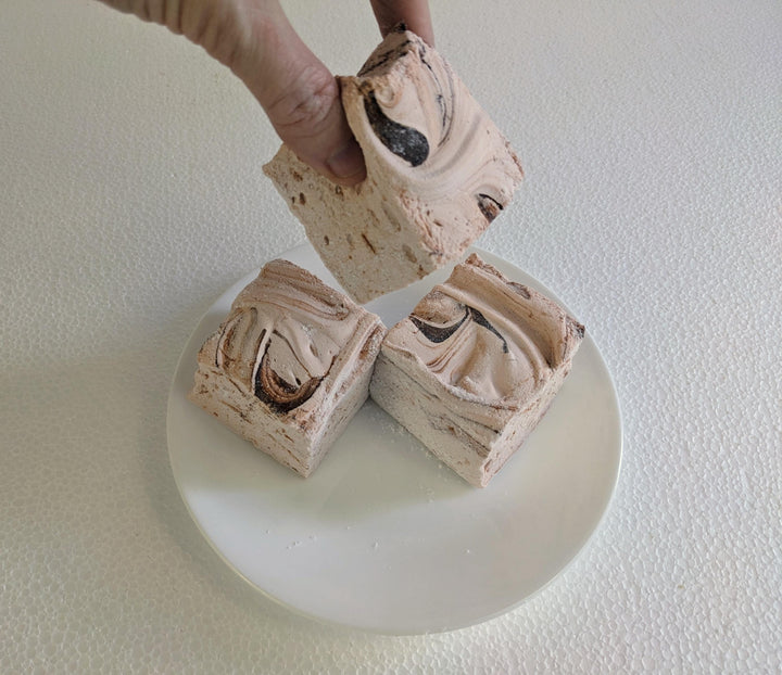 Three handcrafted gourmet Dulce De Leche marshmallows, neatly placed on a white plate and displayed against a plain white background. A hand is shown picking up one of the marshmallows.