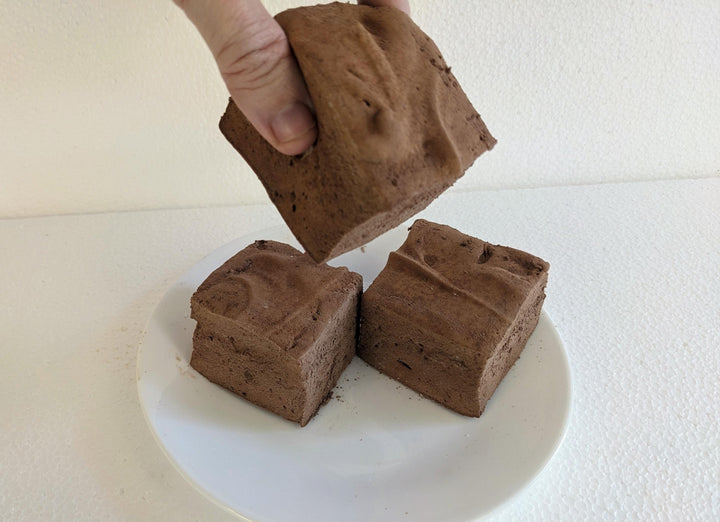 Three Hot chocolate marshmallows on a plate, displayed on a clean, white background. A hand is seen picking up one of the marshmallows.