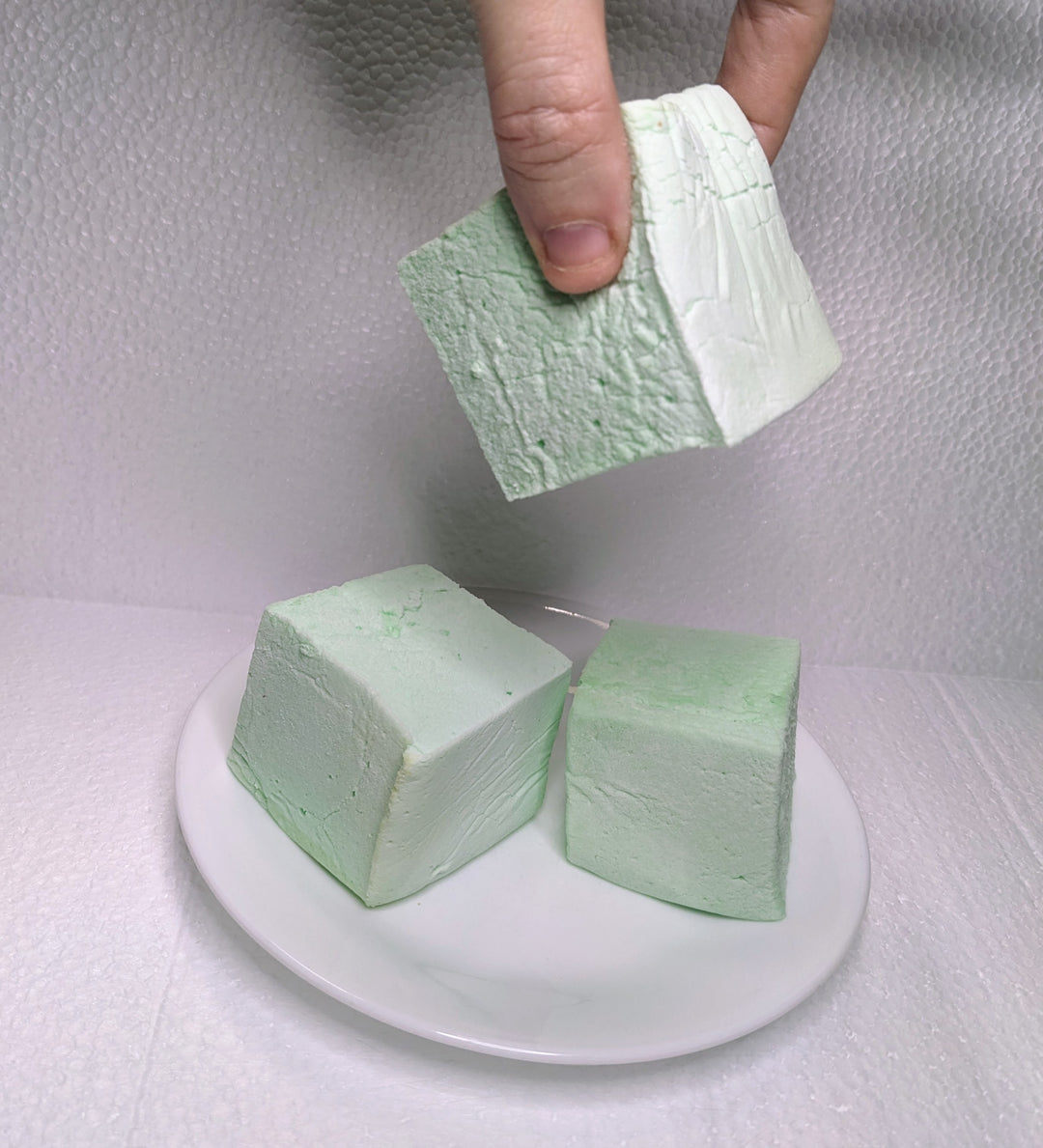 Three mint green marshmallows on a white plate, displayed on a clean, white background. A hand is seen picking up one of the marshmallows.