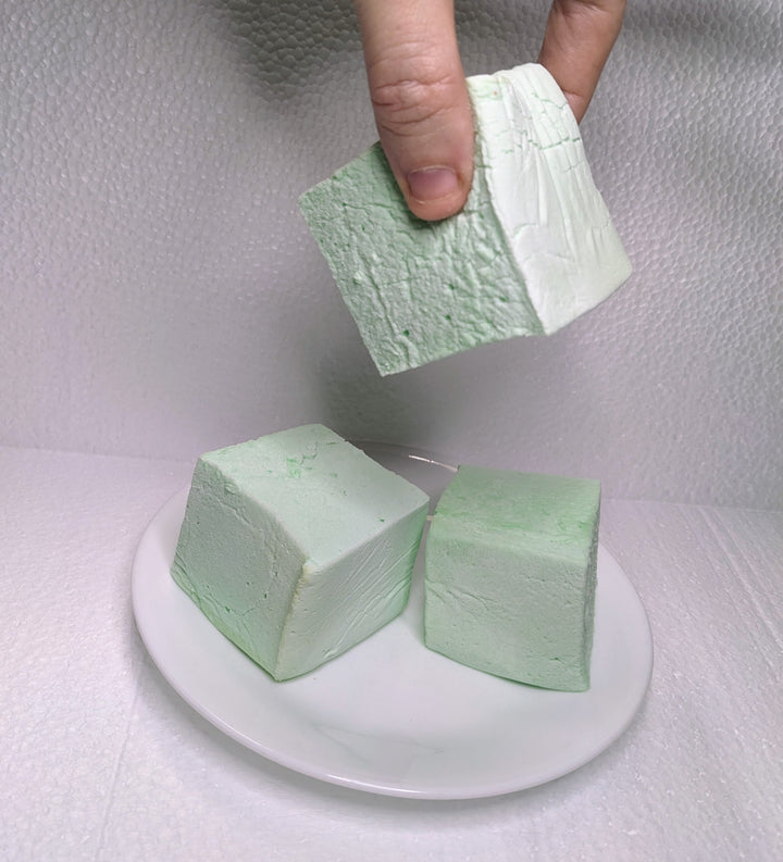Three mint green marshmallows on a white plate, displayed on a clean, white background. A hand is seen picking up one of the marshmallows.