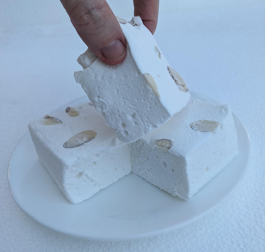 A hand is shown picking up one of three white marshmallows with almond slivers on top, from a white plate with a white background. The marshmallows are cut to approximately 2" x 2" x 1-1.5", showcasing their golden brown, toasted exterior. The white plate and background emphasize the artisanal quality and delightful appearance of the marshmallows.