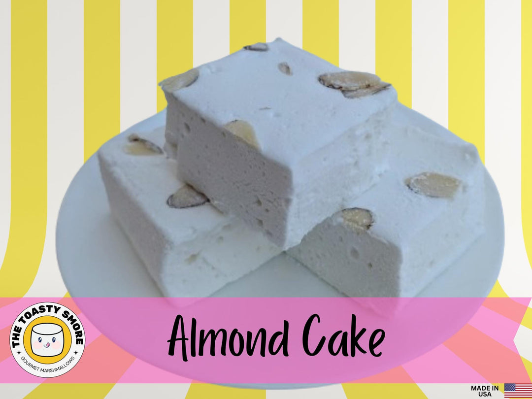 A plate of Handcrafted Almond Cake Flavored Gourmet Marshmallows displayed on a yellow and white striped background. The marshmallows are cut to approximately 2" x 2" x 1.5+", showcasing their golden brown, toasted exterior. The scene features the Toasty Smore logo prominently, along with a "Made in USA" flag, emphasizing the artisanal and local craftsmanship of the product.