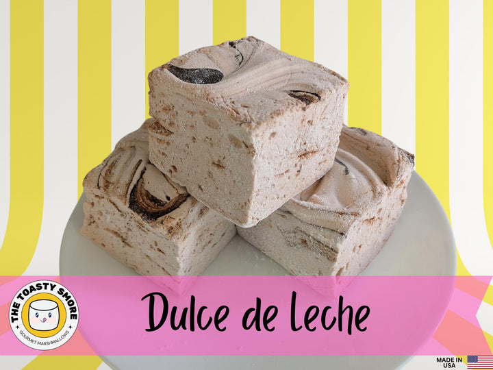 Three handcrafted gourmet Dulce De Leche marshmallows on a white plate, displayed against a yellow and white striped background. The marshmallows are approximately 2" x 2" x 2" each and made from gelatin. The image features The Toasty Smore logo and a "Made in USA" flag.