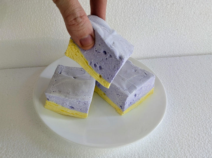 A plate of three handcrafted gourmet lavender & honey marshmallows displayed on a clean white background. The image showcases their soft, fluffy texture and delicate floral flavor. A hand is shown picking up one of the marshmallows for a squeeze test, highlighting its quality and texture.
