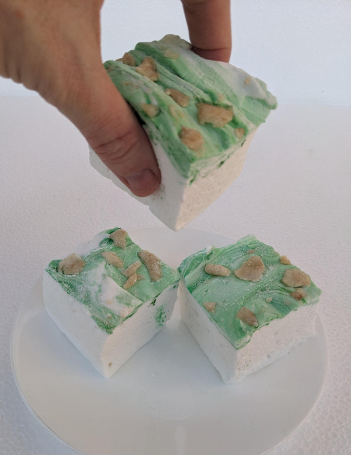 A plate of three handcrafted gourmet pears in wine syrup marshmallows displayed on a clean white background. The marshmallows are approximately 2" x 2" x 1.5" each, arranged on a clean white plate. The image features a hand gently picking up one of the marshmallows, highlighting their soft and fluffy texture.