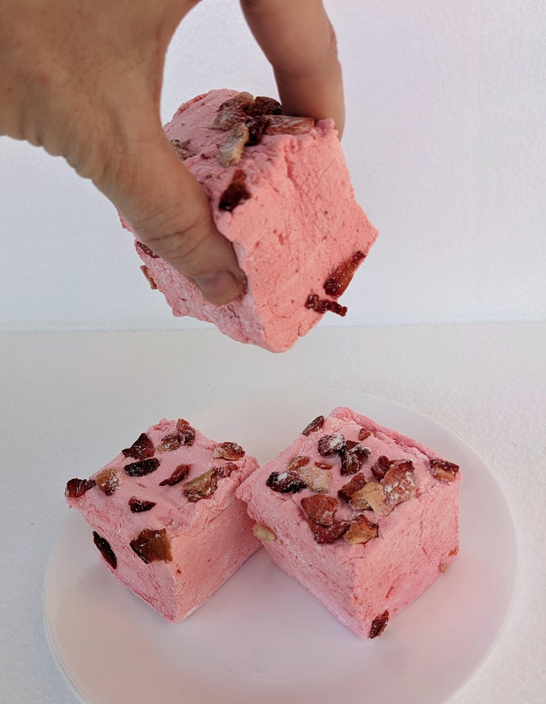 Stack of Three Handcrafted Gourmet Strawberries in White Custard Sauce Marshmallows on a White Background. A hand is gently squeezing one of the marshmallows to show its softness and texture.