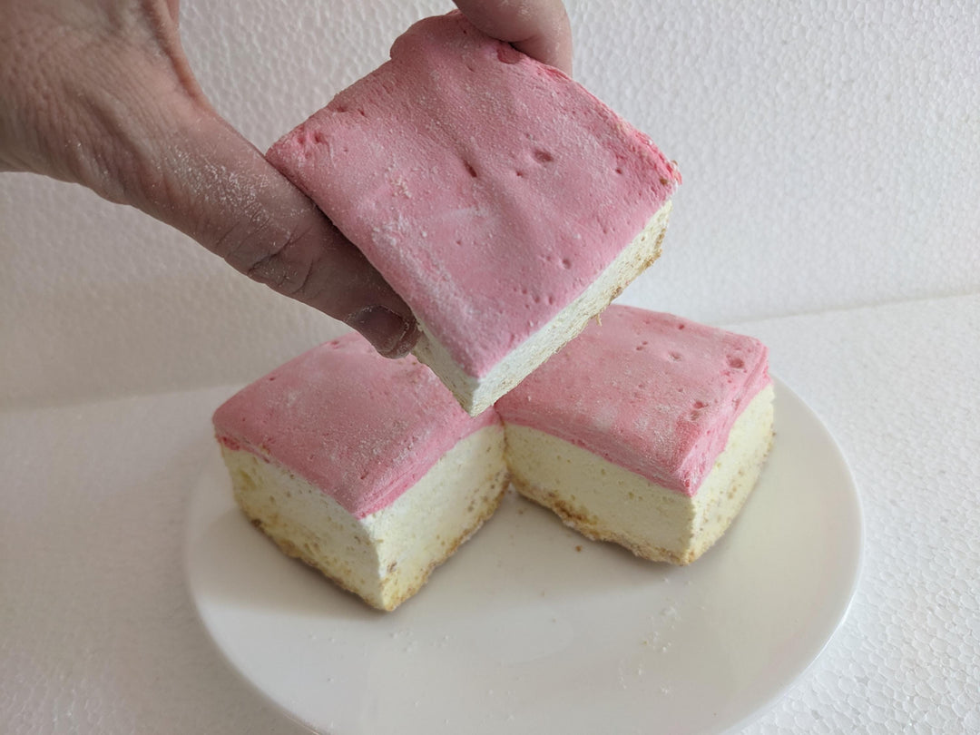 A plate of three handcrafted gourmet strawberry cheesecake marshmallows displayed on a clean white background. The marshmallows are approximately 2" x 2" x 1.5" each, showcasing their soft, fluffy texture and rich cheesecake flavor with strawberry accents. The image features a hand gently squeezing one of the marshmallows, highlighting its soft and fluffy texture.