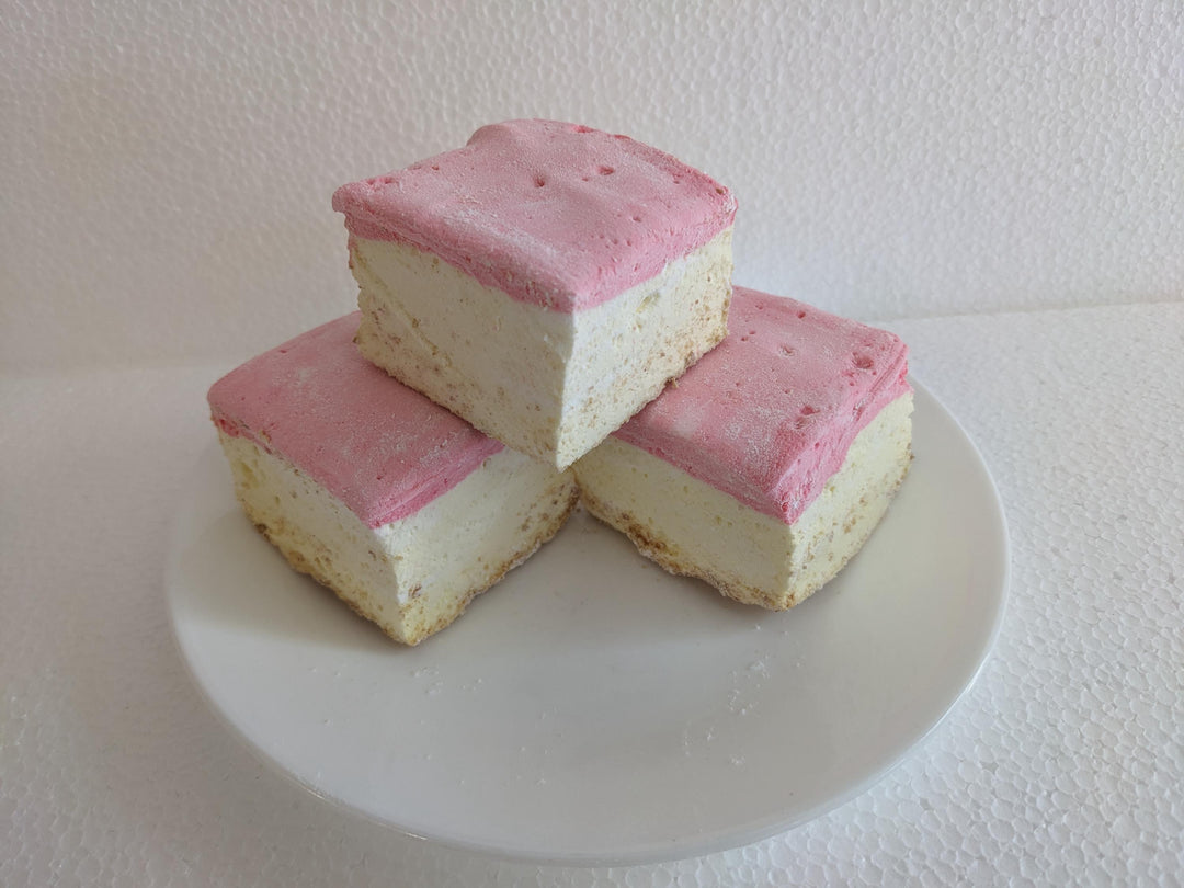 A plate of three handcrafted gourmet strawberry cheesecake marshmallows displayed on a clean white background. The marshmallows are approximately 2" x 2" x 2" each, showcasing their soft, fluffy texture and rich cheesecake flavor with strawberry accents.