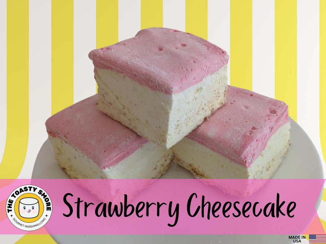 A plate of three handcrafted gourmet strawberry cheesecake marshmallows displayed on a vibrant yellow and white striped background. The marshmallows are approximately 2" x 2" x 2" each, showcasing their soft, fluffy texture and rich cheesecake flavor with strawberry accents. The text prominently features The Toasty Smore logo and a "Made in USA" flag, emphasizing the artisanal quality and craftsmanship.