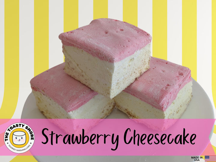 A plate of three handcrafted gourmet strawberry cheesecake marshmallows displayed on a vibrant yellow and white striped background. The marshmallows are approximately 2" x 2" x 2" each, showcasing their soft, fluffy texture and rich cheesecake flavor with strawberry accents. The text prominently features The Toasty Smore logo and a "Made in USA" flag, emphasizing the artisanal quality and craftsmanship.