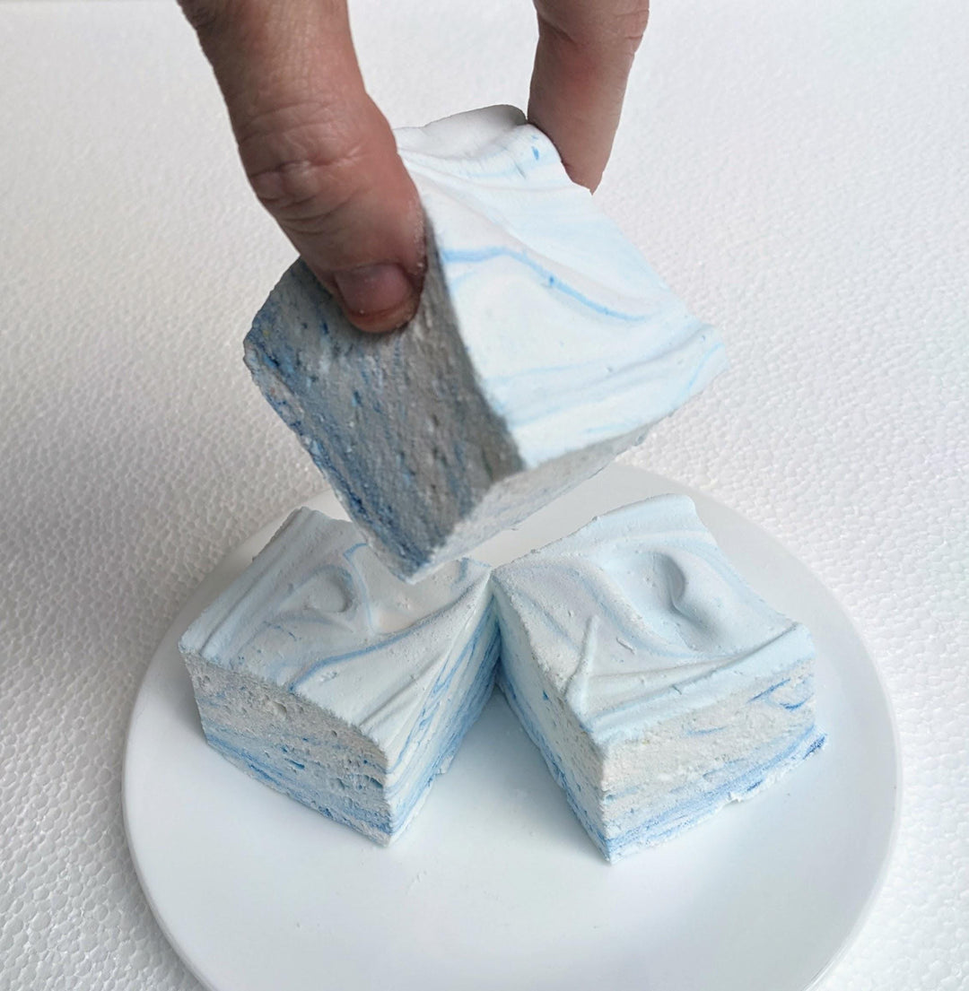 A hand is shown picking up one of three Gourmet Handcrafted Junket with Blueberries Marshmallows from a white plate, highlighting the size and texture of the marshmallow. The white plate and white background provide a clean and simple backdrop, emphasizing the delicious and artisanal quality of the marshmallows.