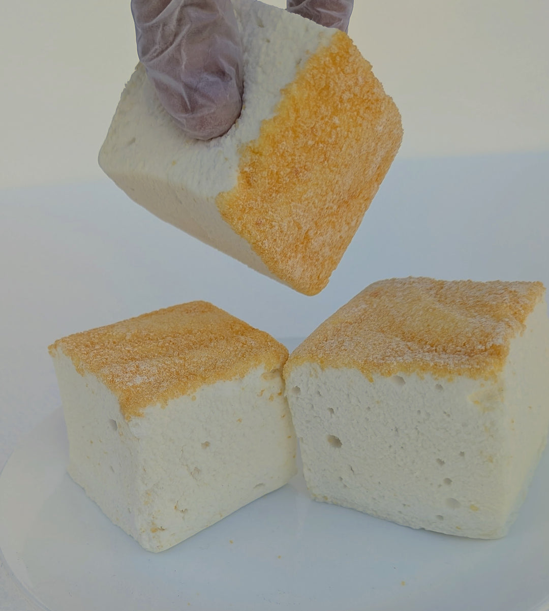 A gloved hand lifting a fluffy Piña Colada marshmallow with toasted top above two more pieces, made with beef gelatin and organic marshmallow root; clean‑label, corn‑syrup free real marshmallows.