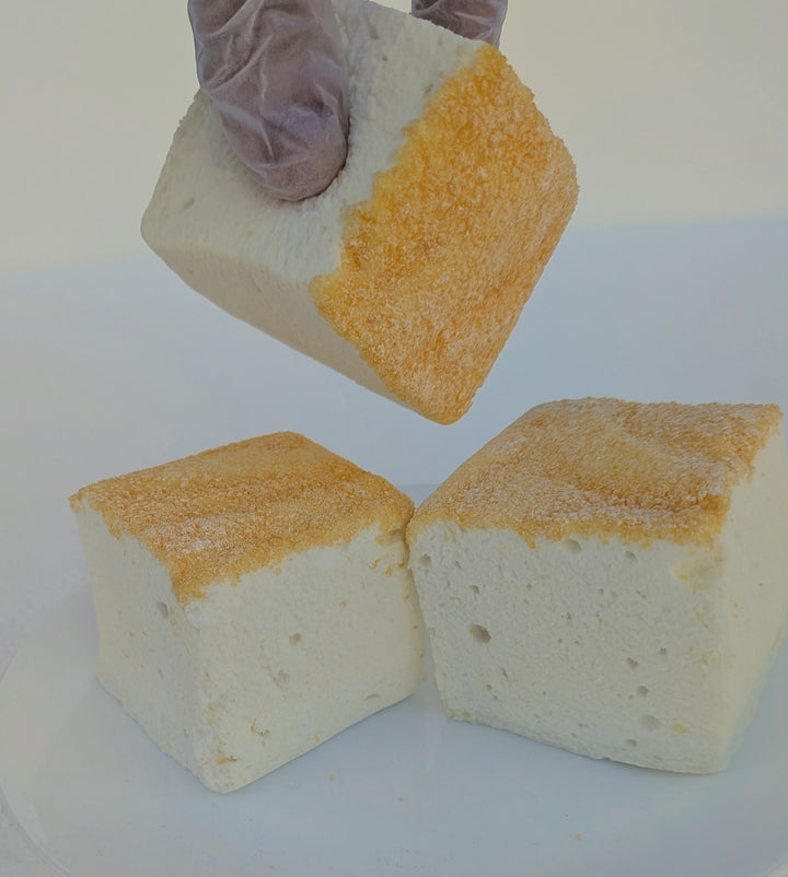 A gloved hand lifting a fluffy Piña Colada marshmallow with toasted top above two more pieces, made with beef gelatin and organic marshmallow root; clean‑label, corn‑syrup free real marshmallows.