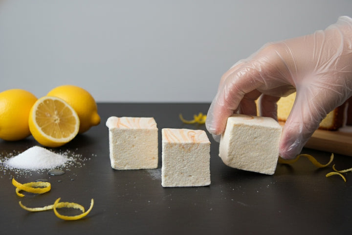 A gloved hand lifts one of three Lemon Pound Cake marshmallows by The Toasty S’more, topped with lemon zest and styled on a dark surface with fresh lemons and a sliced pound cake in the background. Handcrafted, corn-syrup free, and made with beef gelatin and organic marshmallow root for a clean-label indulgence.
