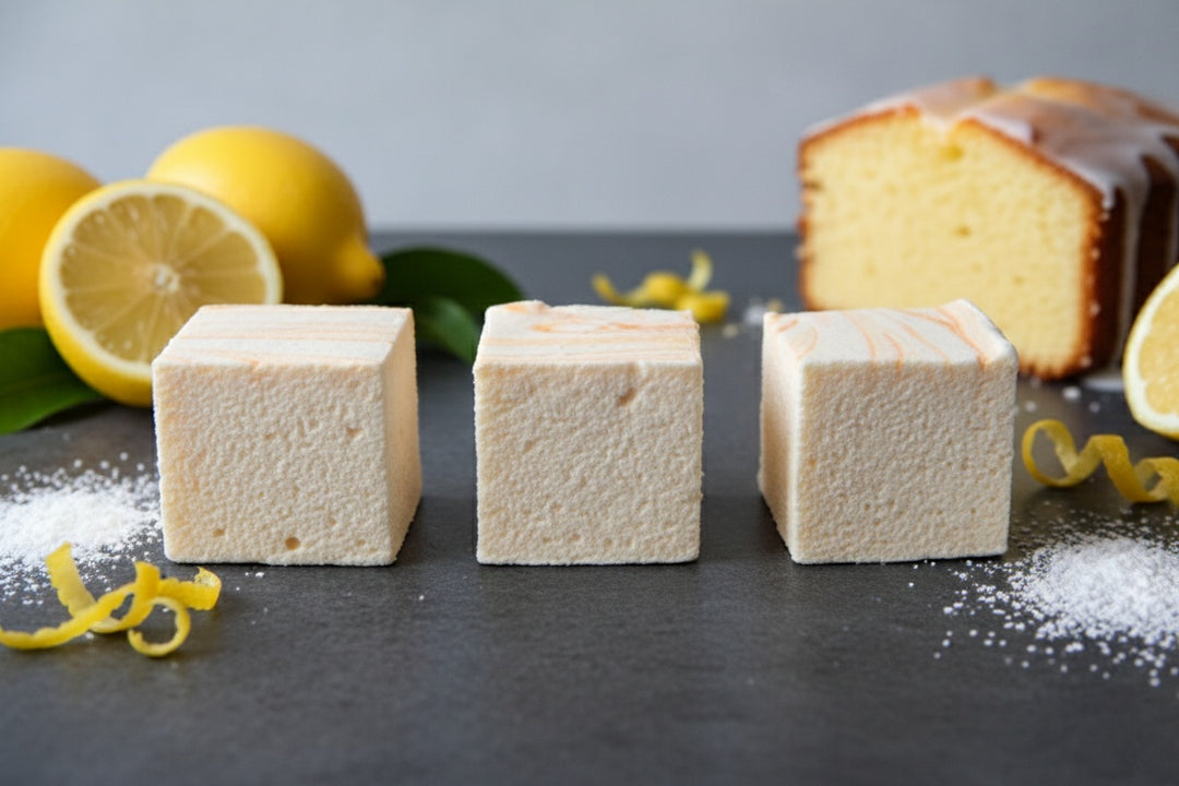 Three handcrafted Lemon Pound Cake marshmallows by The Toasty S’more topped with lemon zest on a dark surface, styled with fresh lemons and a pound cake in the background. Made with beef gelatin and organic marshmallow root for a clean-label, corn-syrup free treat.
