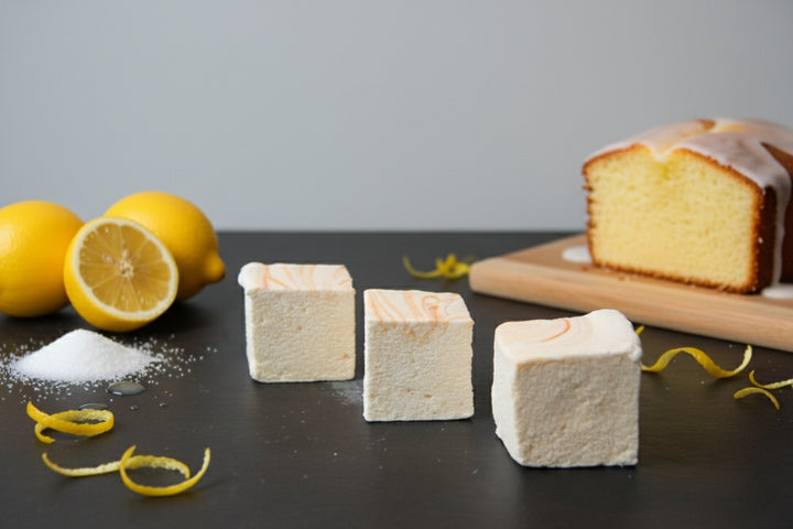Three handcrafted Lemon Pound Cake marshmallows by The Toasty S’more topped with lemon zest, styled on a dark surface with fresh lemons and a sliced pound cake in the background. Made with beef gelatin and organic marshmallow root for a corn-syrup free, clean-label treat.