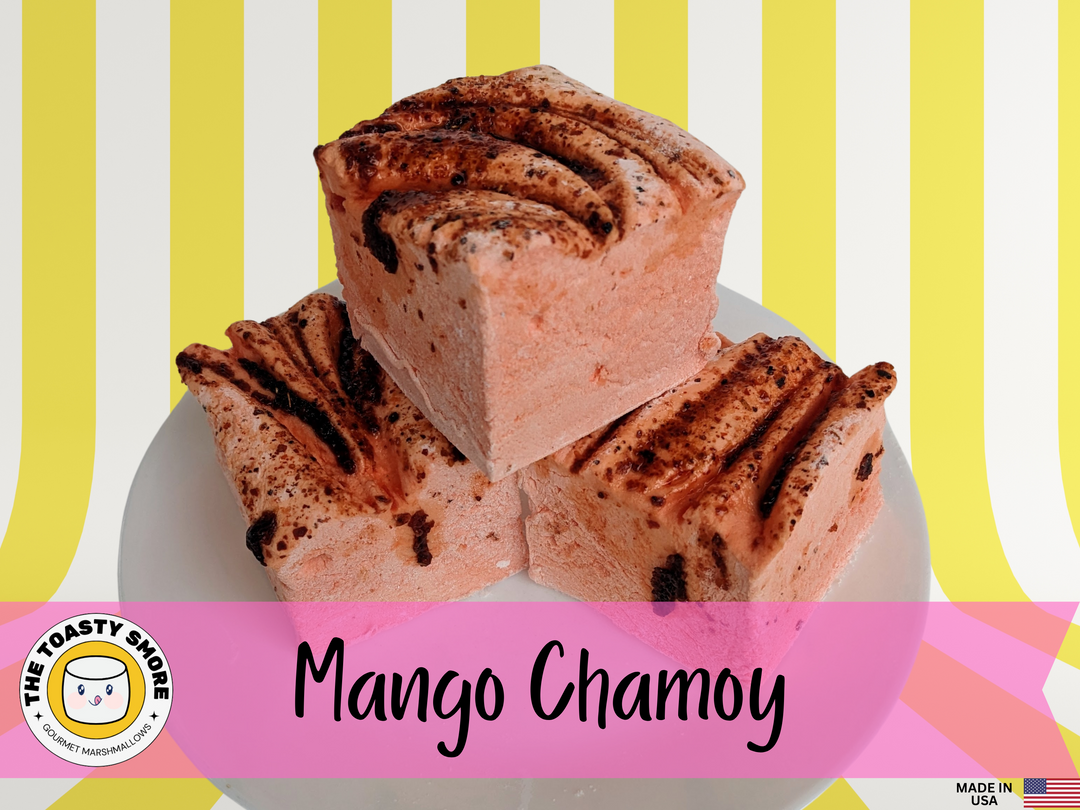 Three vibrant orange mango chamoy marshmallows on a white plate, set against a yellow and white striped background. The marshmallows are displayed with The Toasty Smore logo and a "Made in USA" flag.