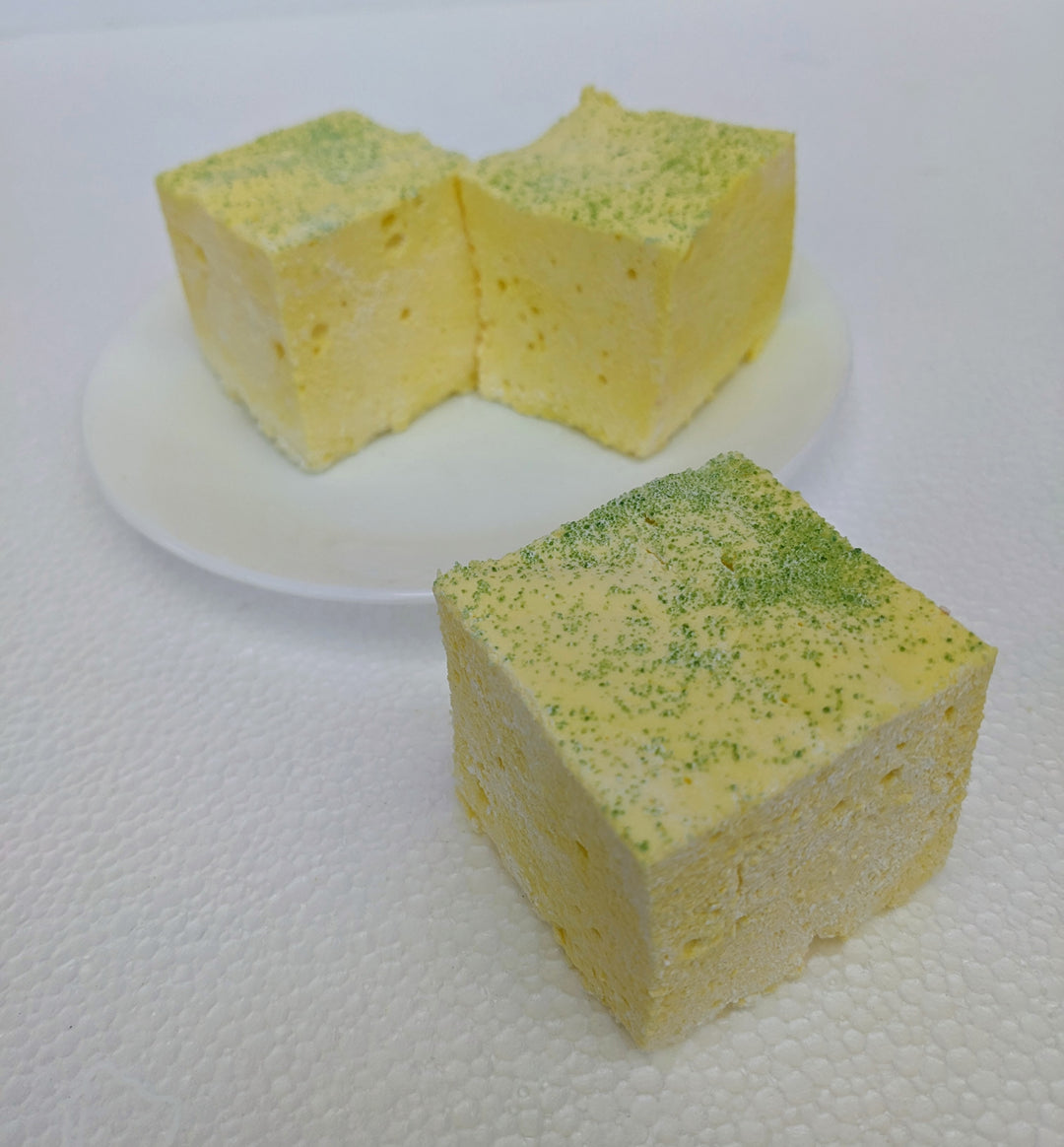 A close-up view of three pineapple-inspired gourmet marshmallows, handcrafted and made in the USA by The Toasty Smore, arranged on a white plate with one marshmallow in front and two in the back. Each marshmallow is yellow with a green sugar topping, resembling a pineapple. Their soft, fluffy texture contrasts with the sparkling sugar, highlighting their artisanal quality. The arrangement adds depth to the image, showcasing the marshmallows as a vibrant tropical treat.