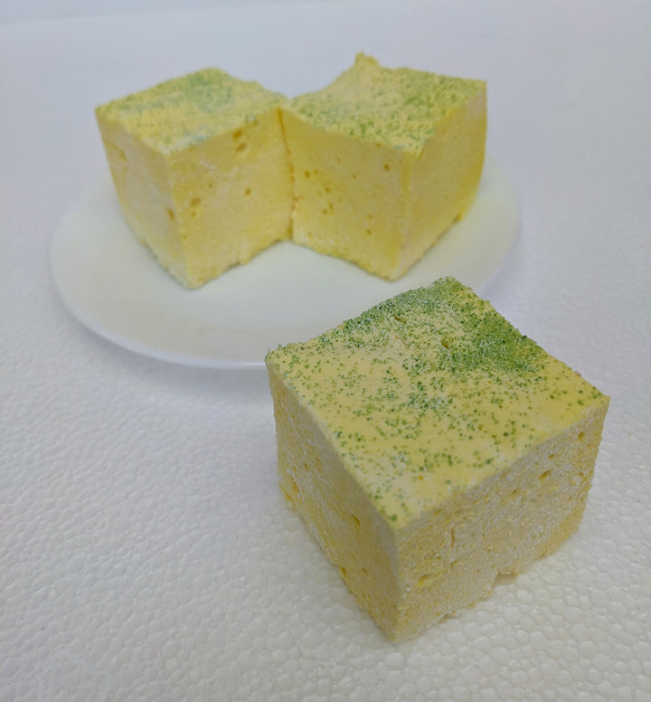A close-up view of three pineapple-inspired gourmet marshmallows, handcrafted and made in the USA by The Toasty Smore, arranged on a white plate with one marshmallow in front and two in the back. Each marshmallow is yellow with a green sugar topping, resembling a pineapple. Their soft, fluffy texture contrasts with the sparkling sugar, highlighting their artisanal quality. The arrangement adds depth to the image, showcasing the marshmallows as a vibrant tropical treat.