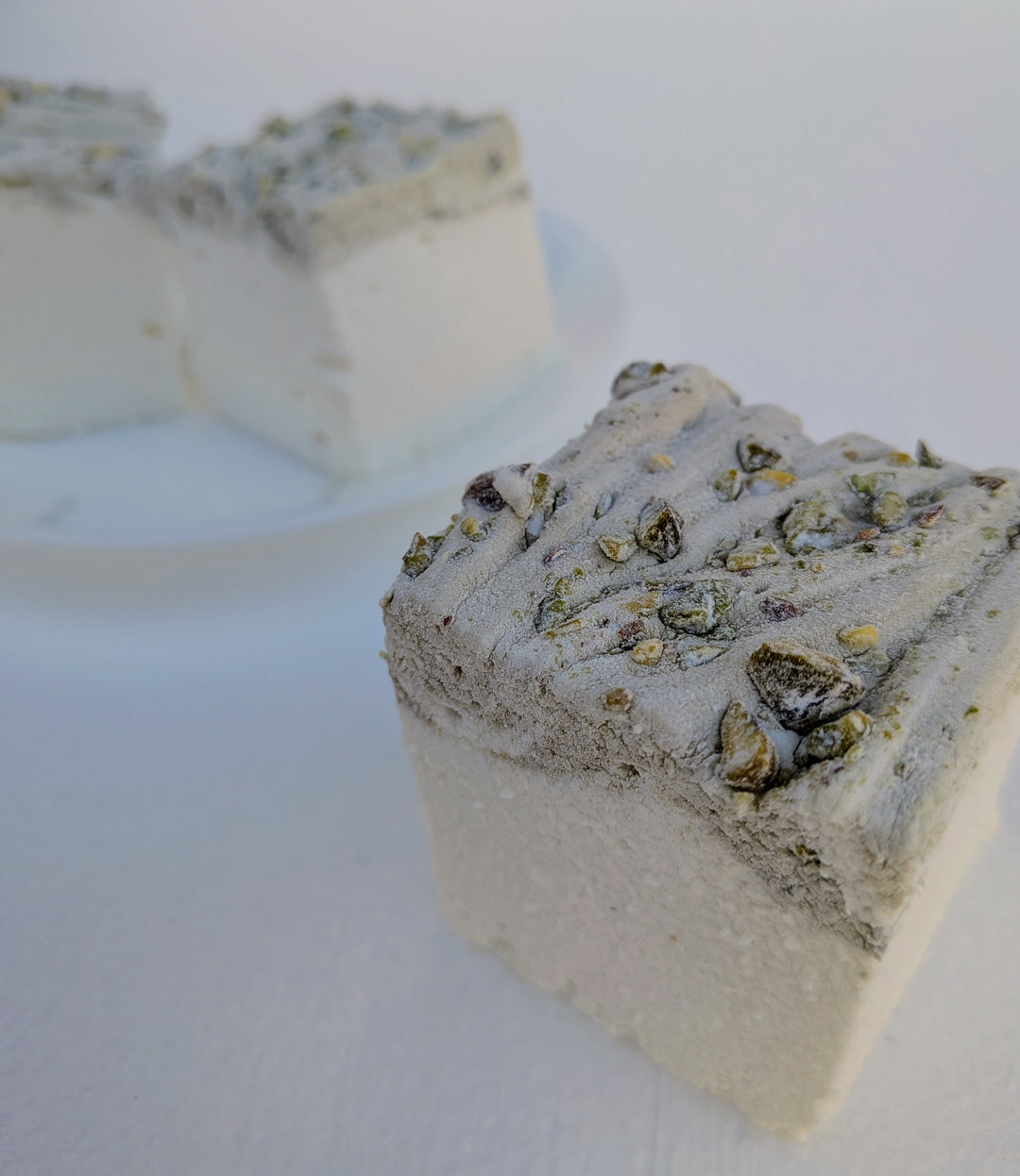 Close‑up of a pistachio marshmallow made with beef gelatin and organic marshmallow root, showing a pistachio‑studded top with two more pieces blurred on a white plate behind it. Clean‑label, gluten‑free, corn syrup‑free real marshmallows photographed in a detailed food shot.