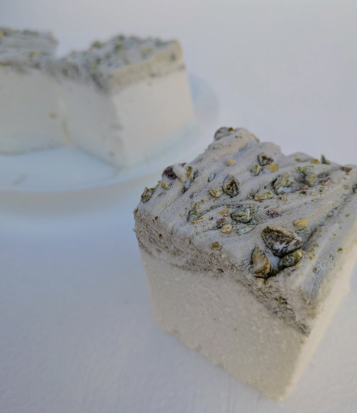 Close‑up of a pistachio marshmallow made with beef gelatin and organic marshmallow root, showing a pistachio‑studded top with two more pieces blurred on a white plate behind it. Clean‑label, gluten‑free, corn syrup‑free real marshmallows photographed in a detailed food shot.