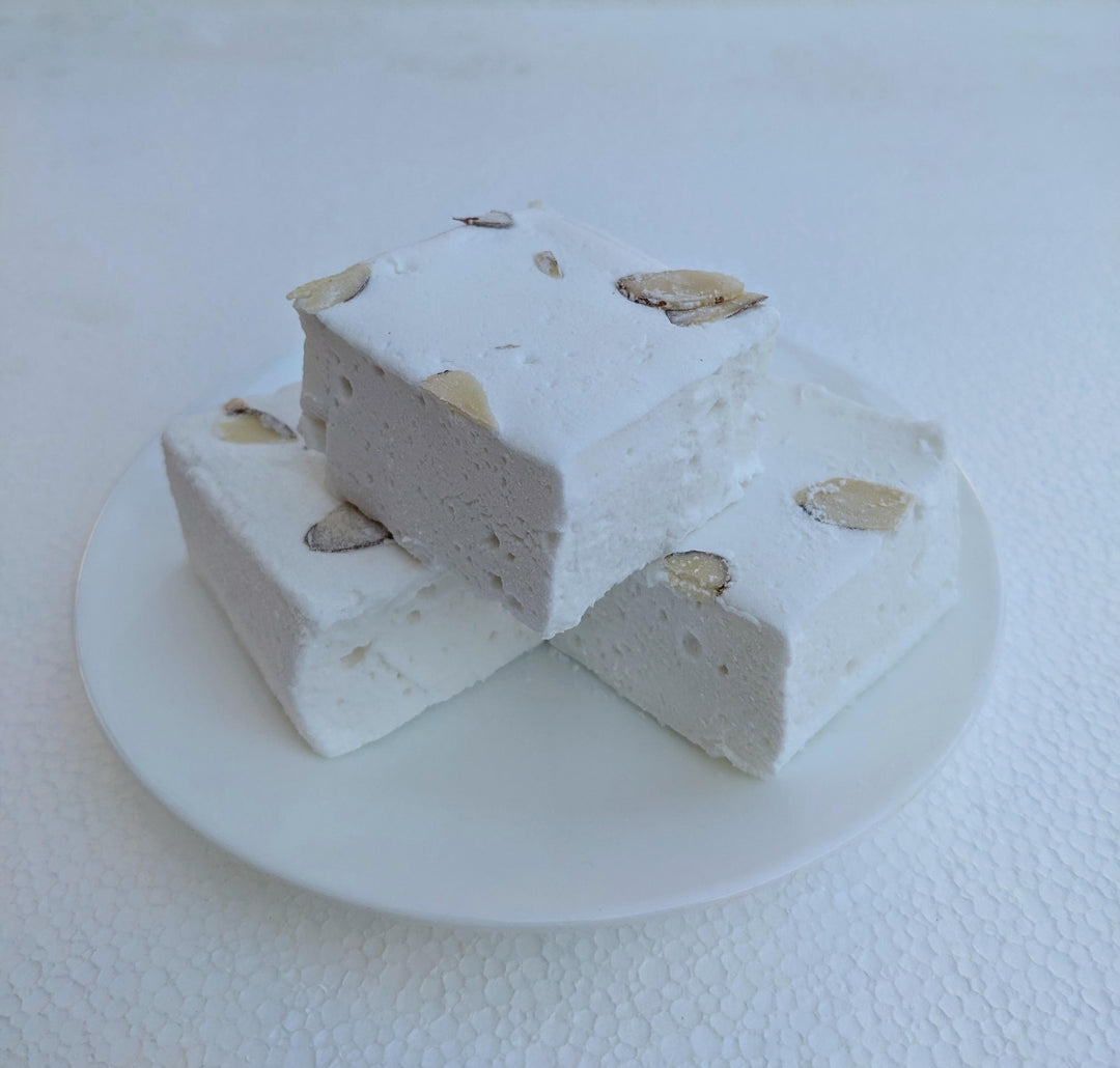 A plate of three almond cake marshmallows displayed on a clean, white plate with a white background. The marshmallows are cut to approximately 2" x 2" x 1.5+", showcasing their white exterior with almond slivers on top. The simplicity of the white plate and background emphasizes the artisanal quality and delightful appearance of the marshmallows.