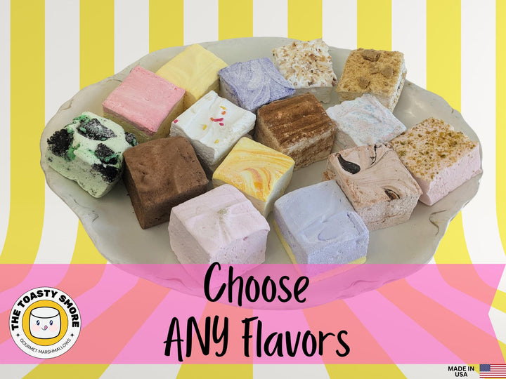 A plate featuring various flavors and colors of gourmet marshmallows by The Toasty Smore. The vibrant marshmallows are displayed on a yellow and white striped background, with a "Made in USA" flag prominently visible. The assortment showcases a delightful range of textures and hues, emphasizing the artisanal craftsmanship of the marshmallows.