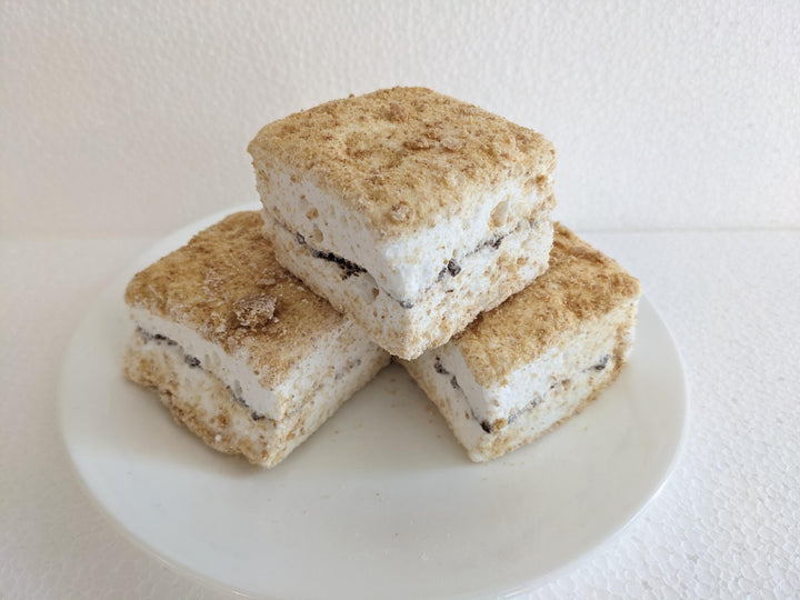 Three s’mores marshmallows layered with graham crumbs and chocolate pieces, stacked on a white plate.