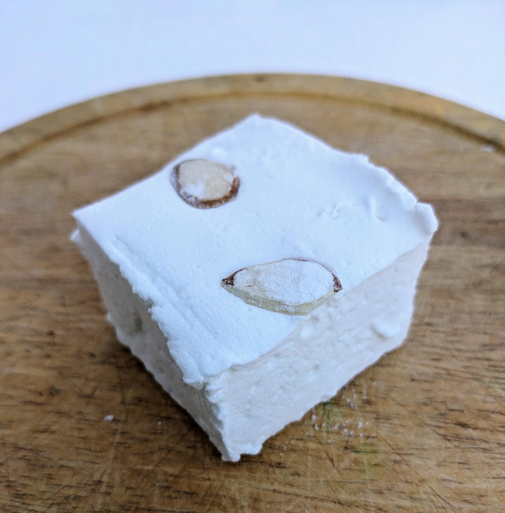 A single almond cake marshmallow displayed on a wooden cutting board. The marshmallow is cut to approximately 2" x 2" x 1.5+", showcasing its pure white exterior. The wooden cutting board provides a rustic and appealing backdrop, emphasizing the artisanal quality of the marshmallow.